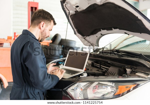 Mid adult auto repair worker using laptop to\
detect malfunction in car at\
garage