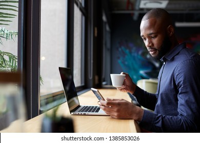 Mid adult black male creative sits by window having coffee, using a laptop and smartphone, side view
