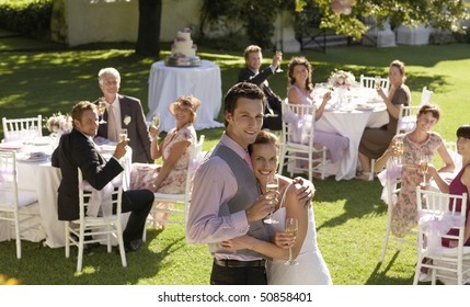 Mid adult bride and groom in garden among wedding guests, holding wineglasses, embracing
