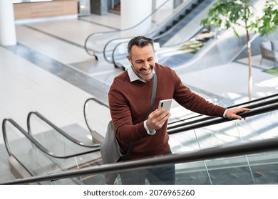 Mid adult business man smiling while reading message on smartphone at airport. Successful senior casual man going up the escalator using smart phone while going to work. Businessman doing video call.