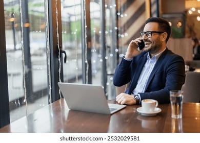 A mid adult Caucasian businessman multitasks with a laptop and smartphone, dressed in smart attire, at a café table by the window, exuding productivity in a casual setting. - Powered by Shutterstock