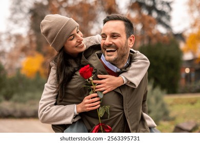 Mid adult Caucasian male playfully carries his partner in a public park on Valentine's Day. She holds a red rose, both dressed warmly in casual winter attire, creating a joyful and romantic scene. - Powered by Shutterstock