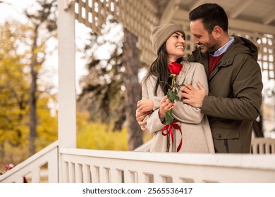 Mid adult Caucasian male presents a red rose to his girlfriend in a charming pavilion for Valentine's Day. The couple  enjoys a joyful moment. Perfect setting enhances romantic gesture. - Powered by Shutterstock