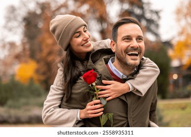 Mid adult Caucasian man piggyback carries smiling woman in park. Both wear casual attire; he sports a jacket, she holds a rose. Outdoors setting reflects seasonal romance.  - Powered by Shutterstock