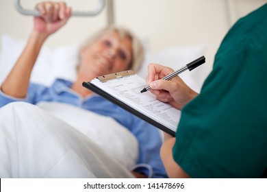 Mid Adult Female Nurse Writing On Clipboard While Looking At Patient In Hospital