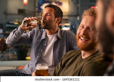 Mid adult friends having fun drinking cold beer sitting on couch at bar. Relaxed man drinking a bottle of beer in a pub while friends having conversation. Three happy young men talking. - Powered by Shutterstock