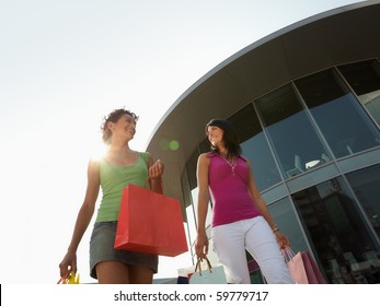 mid adult italian woman and hispanic woman carrying shopping bags out of shopping center at sunset. Horizontal shape, low angle view, copy space