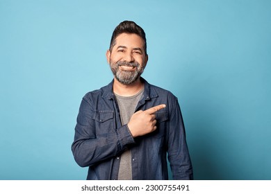 Mid adult latin man smiling pointing to the side showing copy blank space, isolated over blue background. Indoor studio shot. Advertisement, presentation concept. Copy space. - Shutterstock ID 2300755491