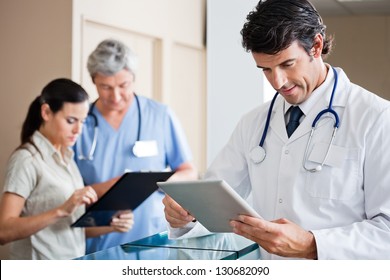 Mid adult male doctor holding digital tablet with colleague and receptionist standing in background - Powered by Shutterstock