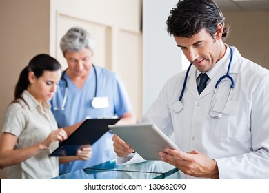 Mid Adult Male Doctor Holding Digital Tablet With Colleague And Receptionist Standing In Background