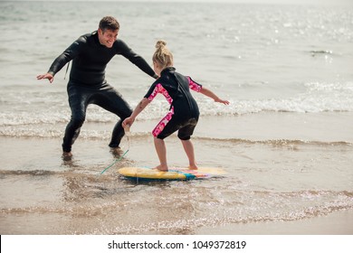 Mid Adult Male Teaching A Little Girl How To Surf At The Beach.