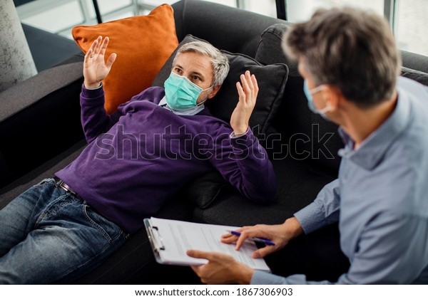 Mid adult man communicating with his therapist\
while lying down on psychiatrist\'s couch and wearing face mask due\
to COVID-19 pandemic. 