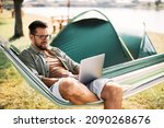 Mid adult man relaxing in hammock while working on laptop during camping day in nature.