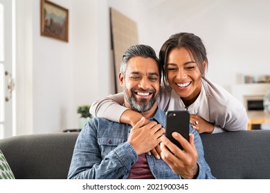 Mid Adult Man Relaxing On Sofa And Showing New App To African American Wife On Cellphone. Middle Eastern Man And Woman Sitting On Couch At Home And Using Mobile Phone To Do A Video Call With Family.