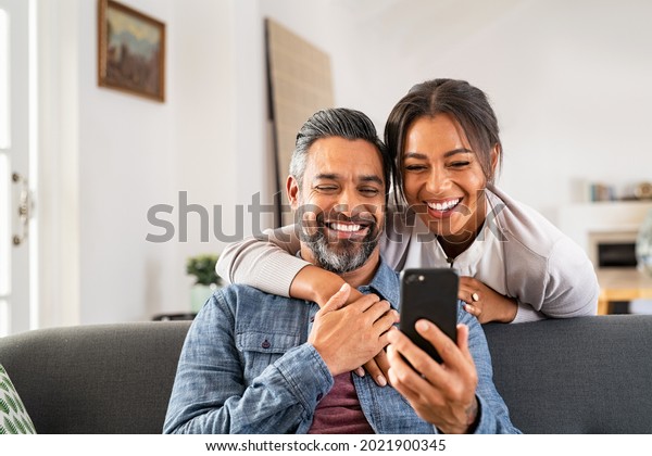 Mid adult man relaxing on sofa and showing new\
app to african american wife on cellphone. Middle eastern man and\
woman sitting on couch at home and using mobile phone to do a video\
call with family.