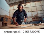 Mid adult man smiling while wearing ear muffs and using power tool on timber in woodworking factory