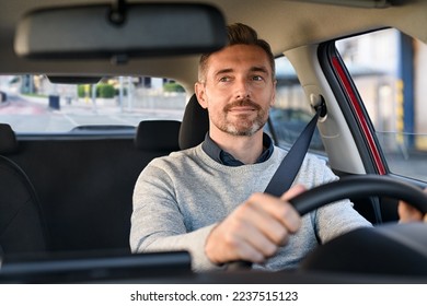Mid adult man smiling while driving car and looking at mirror for reverse. Happy man feeling comfortable sitting on driver seat in his new car. Smiling mature businessman with seat belt on driving. - Shutterstock ID 2237515123
