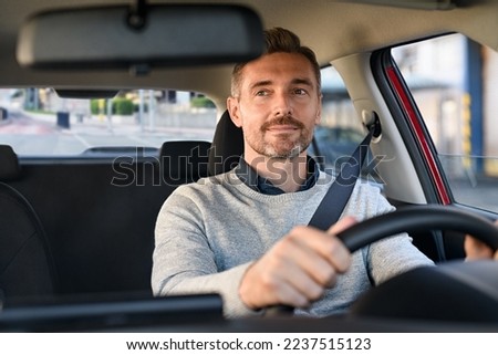 Mid adult man smiling while driving car and looking at mirror for reverse. Happy man feeling comfortable sitting on driver seat in his new car. Smiling mature businessman with seat belt on driving.
