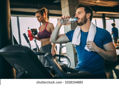 Mid Adult Man Training Hardly On Cycling Machine In Light Spacious Gym. Drinking Water.