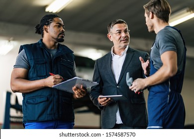 Mid Adult Manager Using Digital Tablet While Talking To Car Mechanics In Auto Repair Shop. 