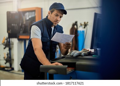 Mid Adult Mechanic Using Computer While Doing Some Paperwork In Auto Repair Shop. 