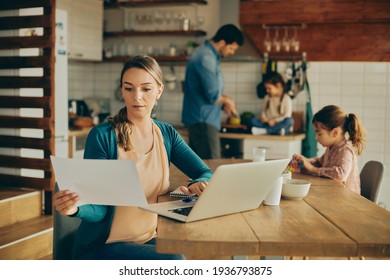 Mid Adult Mother Working On Computer And Examining Reports At Home. Her Husband And Kids Are In The Background. 