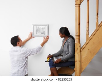 Mid Adult Multiethnic Couple Hanging Pictures To The Wall And Smiling. Horizontal Shape, Copy Space