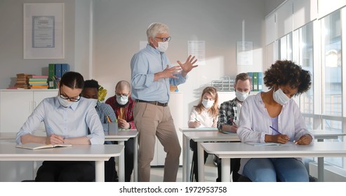 Mid Adult Professor Wearing Face Mask While Talking To A Group Of College Students In Lecture Room. Mature Teacher In Safety Mask Teaching Young People At Adult Education Class