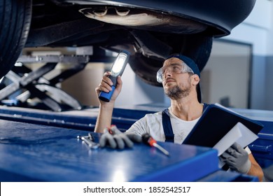 Mid Adult Repairman Using Flashlight While Examining Undercarriage Of A Car In Auto Repair Shop.