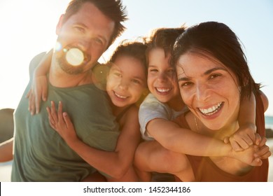 Mid adult white parents piggybacking their kids on a beach, smiling to camera, close up, lens flare