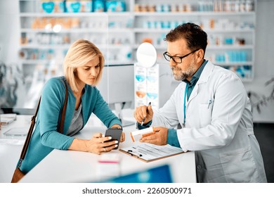 Mid adult woman using cell phone while consulting with her pharmacist in choosing medicine in a pharmacy. - Powered by Shutterstock