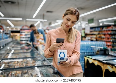 Mid adult woman using smart phone while scanning code on a product in supermarket. 