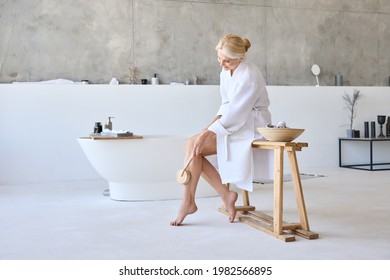 Mid Age Adult 50s Age Mature Older Woman Massaging Legs With Brush Sitting On Bathtub Wearing White Bathrobe In Bathroom. Body Dry Skin Care, Anti Cellulite Massage, Scrub Peeling Exfoliation.