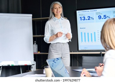 Mid Age Smiling Senior Asian Business Woman Ceo Executive Manager Teacher Presenting To Trainees Interns Income Revenue Data Results On Big Screen In Modern Office On Business Seminar.