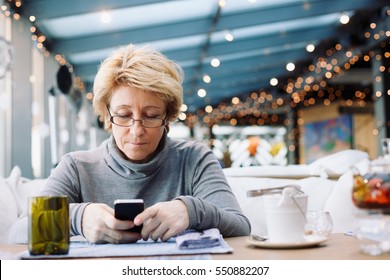 Mid Age Woman With Mobile Phone Sitting At Indoor Cafe Looking For Screen