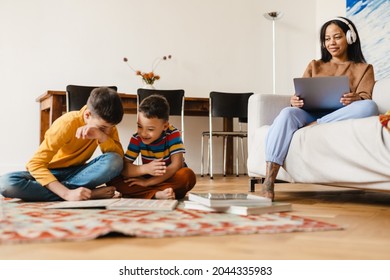 Mid Aged African American Woman Mother Working On Laptop Computer While Her Two Kids Brothers Playing At Home