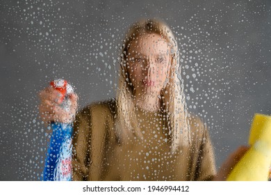 Mid aged blonde woman maid holding bottle of detergent and a rag, spraying glass