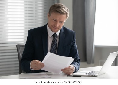 Mid Aged Business Man In Suit Holding Read Document Paper Report Sit At Office Desk, Company Ceo Doing Paperwork Feel Satisfied With Good Work Result Receive Loan Approval Salary Payment Tax Refund