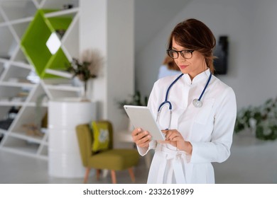 Mid aged female doctor standing at the hospital and holding digital tablet in her hand. Female healthcare worker using touchpad for work.  - Powered by Shutterstock