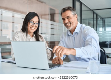 Mid aged Latin male manager mentor teaching young Asian female worker looking at laptop discussing corporate strategy in teamwork, working on computer in office at international team meeting.