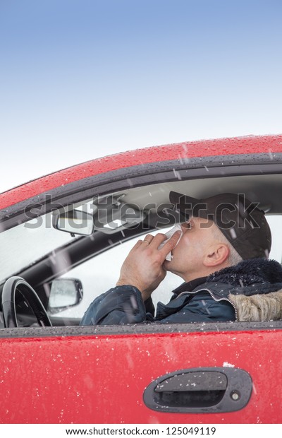 Mid aged\
man with flu symptoms sneezes in a car on snowy  winter day. Using\
handkerchief closes mouth with his\
hands