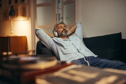 Mid Aged Man Listening Music With Headphones On Phonograph, Relaxed In Sofa At His Home