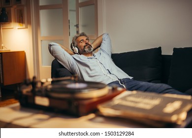 Mann mittleren Alters hört Musik mit Kopfhörern auf einem Plattenspieler, entspannt im Sofa zu Hause