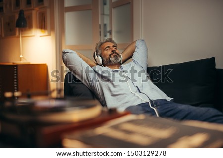mid aged man listening music with headphones on phonograph, relaxed in sofa at his home