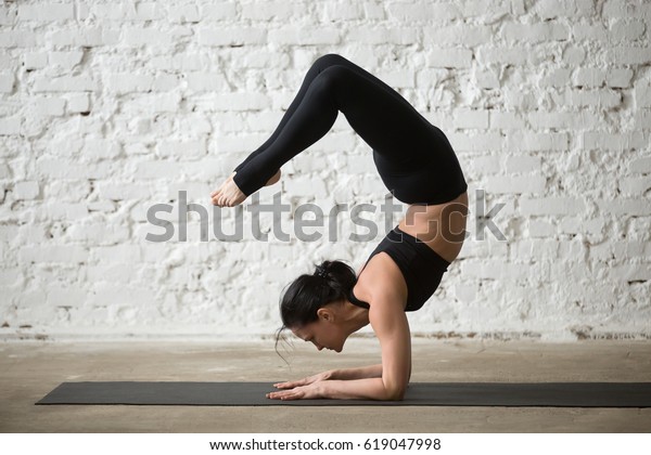 Mid aged yogi attractive woman practicing yoga\
concept, standing in vrischikasana exercise, Scorpion pose, working\
out, wearing sportswear, black tank top and pants, full length,\
white loft background