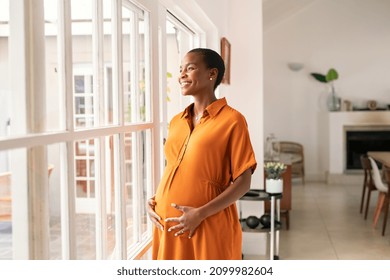 Mid black pregnant woman standing near window at home and thinking about her future family. Smiling african american lady with hands on belly imagine the growth of his baby. Middle aged pregnant woman