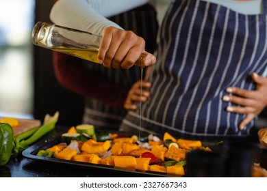 Mid section of biracial lesbian couple embracing and pouring oil on vegetables in kitchen. Cooking, food, healthy lifestyle, lifestyle, relationship, togetherness and domestic life, unaltered. - Powered by Shutterstock