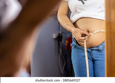 Mid Section Of An Indian Woman Measuring Fat Belly With Tape In Front Of Mirror