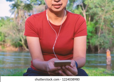 Mid Section Portrait Of An Asian Woman In Red T Shirt Sitting In The Woods With River Having Ear Piece To Listen To Music From Mobile Phone. 