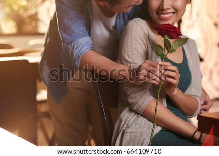 Mid section portrait of man presenting red rose to smiling Asian woman, meeting for date in cafe